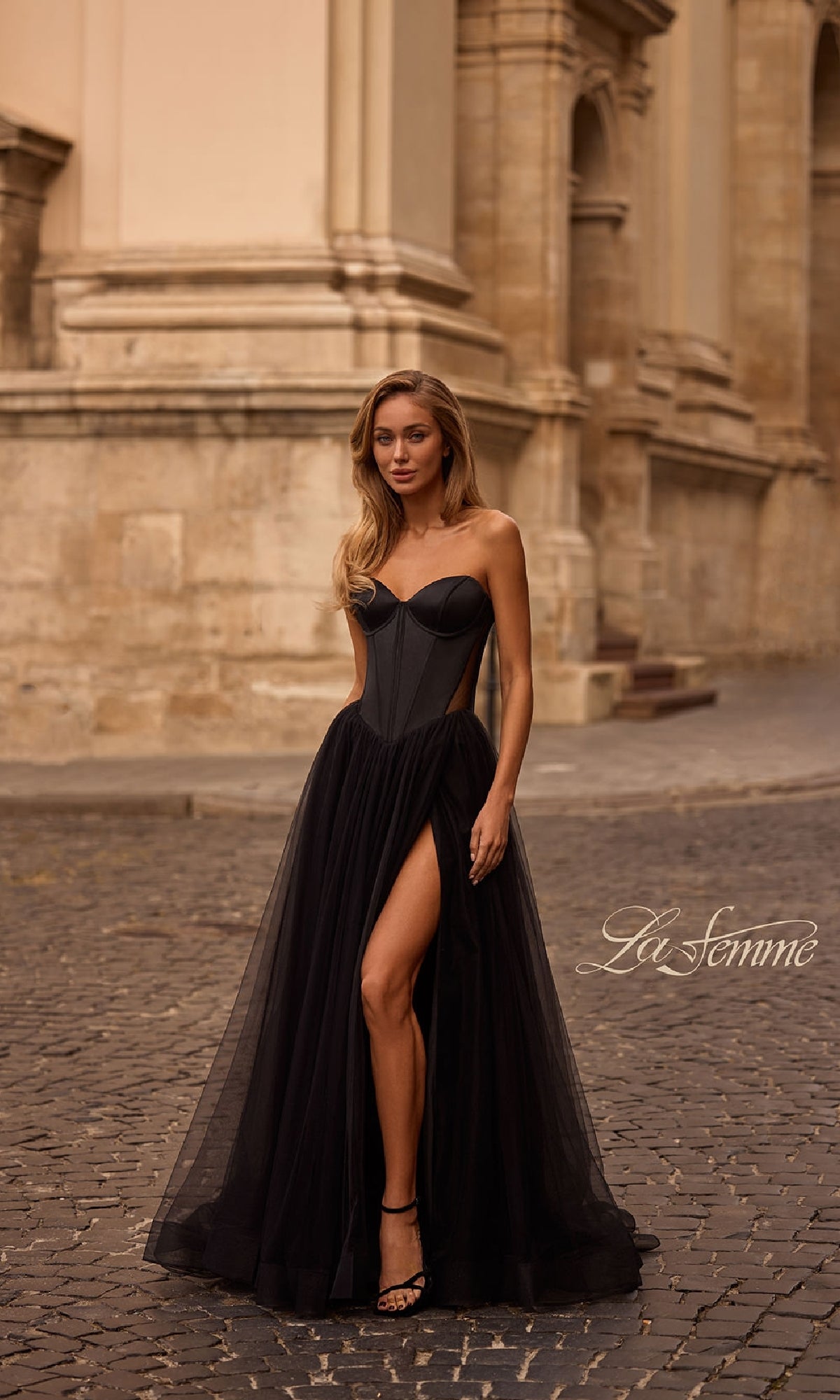 A woman wears the La Femme 32771 black strapless long prom dress with a sweetheart neckline, sheer skirt, and high slit as she stands on a cobblestone street before a historic stone building.